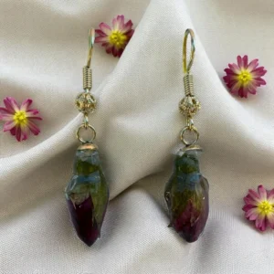 Boucles d’oreilles artisanales dorées avec véritables boutons de rose séchés, montées sur crochets dorés, présentées sur tissu crème avec petites fleurs naturelles – bijou floral fait main par L’Atelier d’Emy-Lys