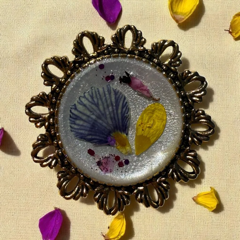 Broche artisanale ronde avec composition florale en pétales de primevère, ficaire et bruyère d’hiver, création unique et poétique.