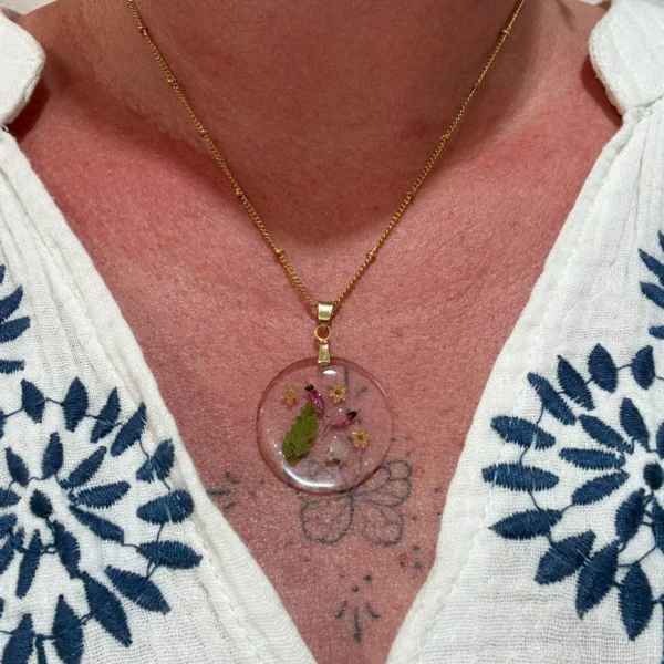 Collier rond doré en résine avec fougère et fleurs naturelles, porté au cou sur chemise blanche à motif fleuri, style bohème.