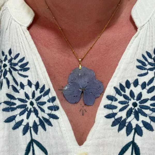Collier doré avec hortensia naturel bleu pâle, bijou poétique et botanique, porté sur décolleté orné d’une blouse brodée.