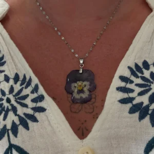 Collier artisanal avec fleur de pensée violette séchée, monté sur chaîne argentée, porté sur peau nue et blouse à fleurs bleues.