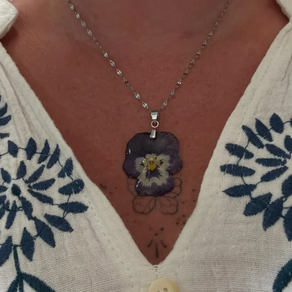 Collier artisanal avec fleur de pensée violette séchée, monté sur chaîne argentée, porté sur peau nue et blouse à fleurs bleues.