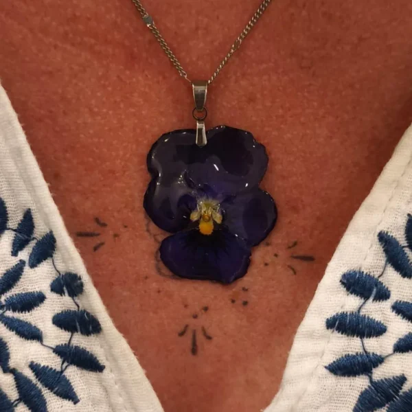 Collier fait main avec pensée naturelle violette, bijou floral en résine porté autour du cou, ambiance artisanale.