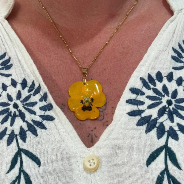 Collier doré avec véritable pensée jaune séchée montée en pendentif floral, porté autour du cou avec blouse brodée – bijou artisanal L’Atelier d’Emy‑Lys