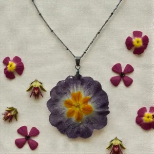 Pendentif fleuri en résine avec primevère violette et centre orange, présenté à plat entouré de fleurs séchées, bijou nature signé L’Atelier d’Emy-Lys.