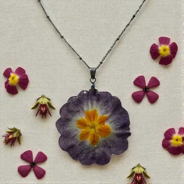 Pendentif fleuri en résine avec primevère violette et centre orange, présenté à plat entouré de fleurs séchées, bijou nature signé L’Atelier d’Emy-Lys.